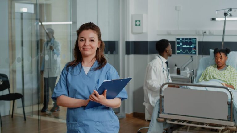 Una chica pelirroja y sonriente, con un pijama de hospital azul hace de celadora o recepcionista sanitaria. Sostiene una carpeta en sus manos. De fondo, un doctor inspecciona a un paciente.